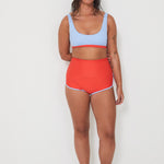 Woman wearing a light blue sports swim bralette and red swim shorts on a white background