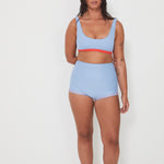Woman wearing a light blue bralette surf top and high-waisted original betty bottom shorts on a white background