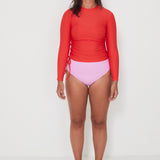 Woman wearing a red long-sleeve  ruched rashguard, salt gypsy swimwear top and pink high-waisted bikini drifter bottoms on a white background, ready for a beach swim