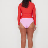 women wearing a red ruched rashguard long-sleeve swim top and pink high-waisted drifter bikini bottom on a white background ready for a surf and swim