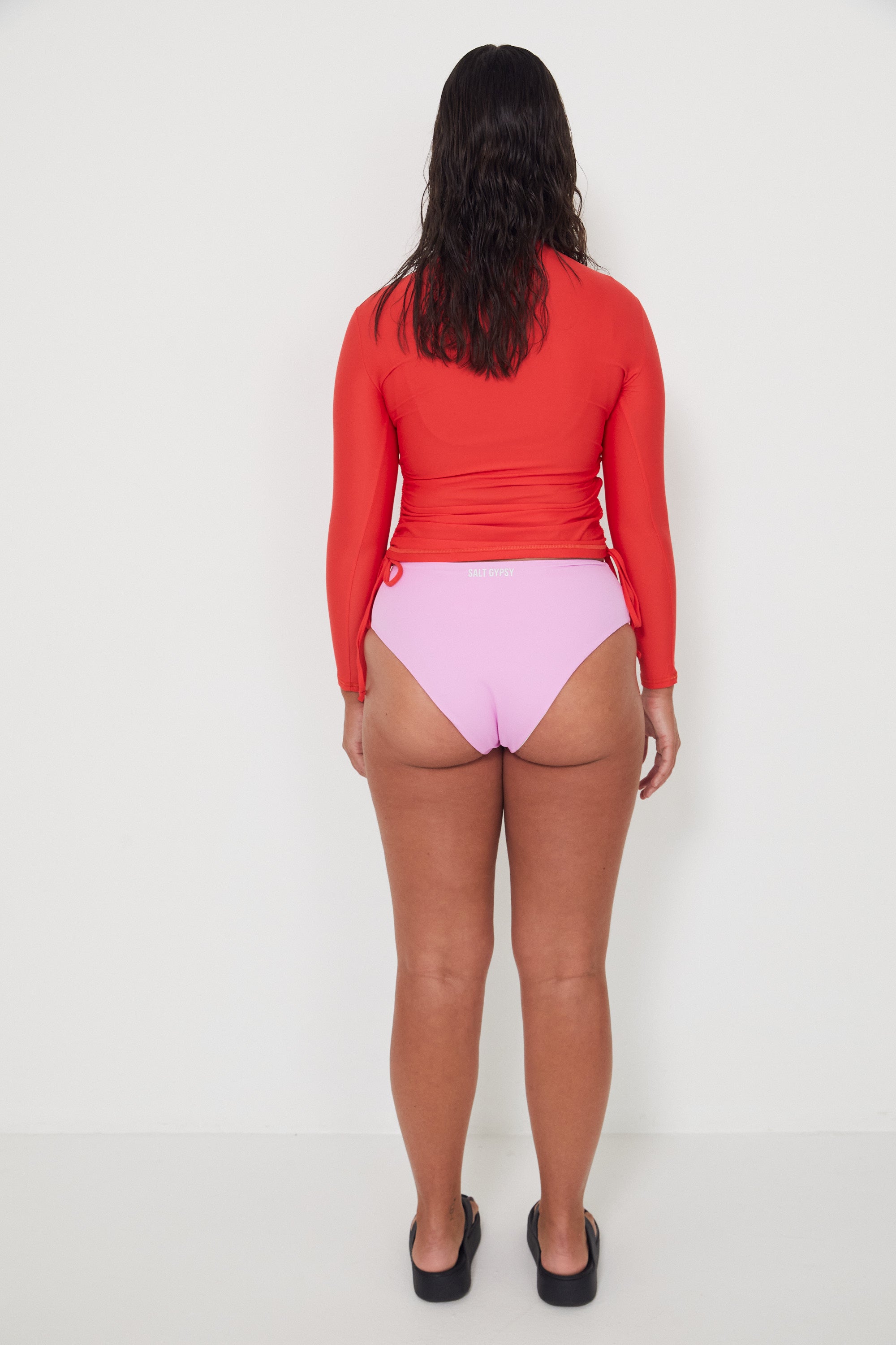 women wearing a red ruched rashguard long-sleeve swim top and pink high-waisted drifter bikini bottom on a white background ready for a surf and swim