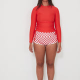 Women wearing a red ruched rashguard long-sleeve swimwear shirt and checkered betty bottom shorts on a white background ready for a surf and swim