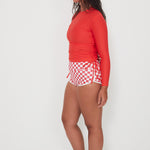 Woman wearing a red long-sleeve ruched rashugard swimwear top and checkered betty bottoms salt gypsy swimwear shorts on a white background ready for a swim at the beach