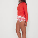 Woman wearing a red long-sleeve ruched rashugard swimwear top and checkered betty bottoms salt gypsy swimwear shorts on a white background ready for a swim at the beach