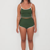 Woman wearing a green tankini surf top and betty bottoms 2.0 swimsuit with white trim against a white background