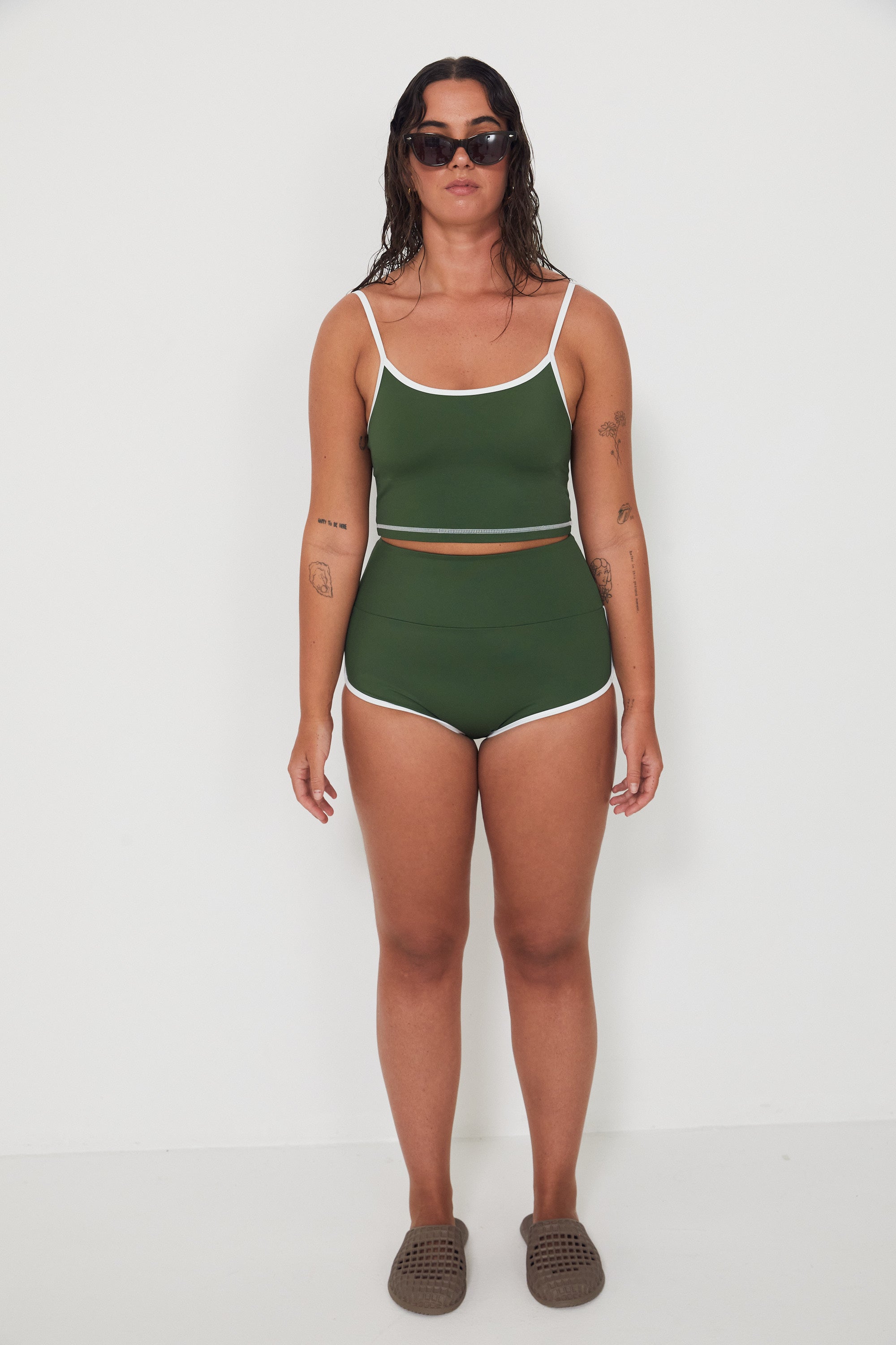 Woman wearing a green tankini surf top and betty bottoms 2.0 swimsuit with white trim against a white background