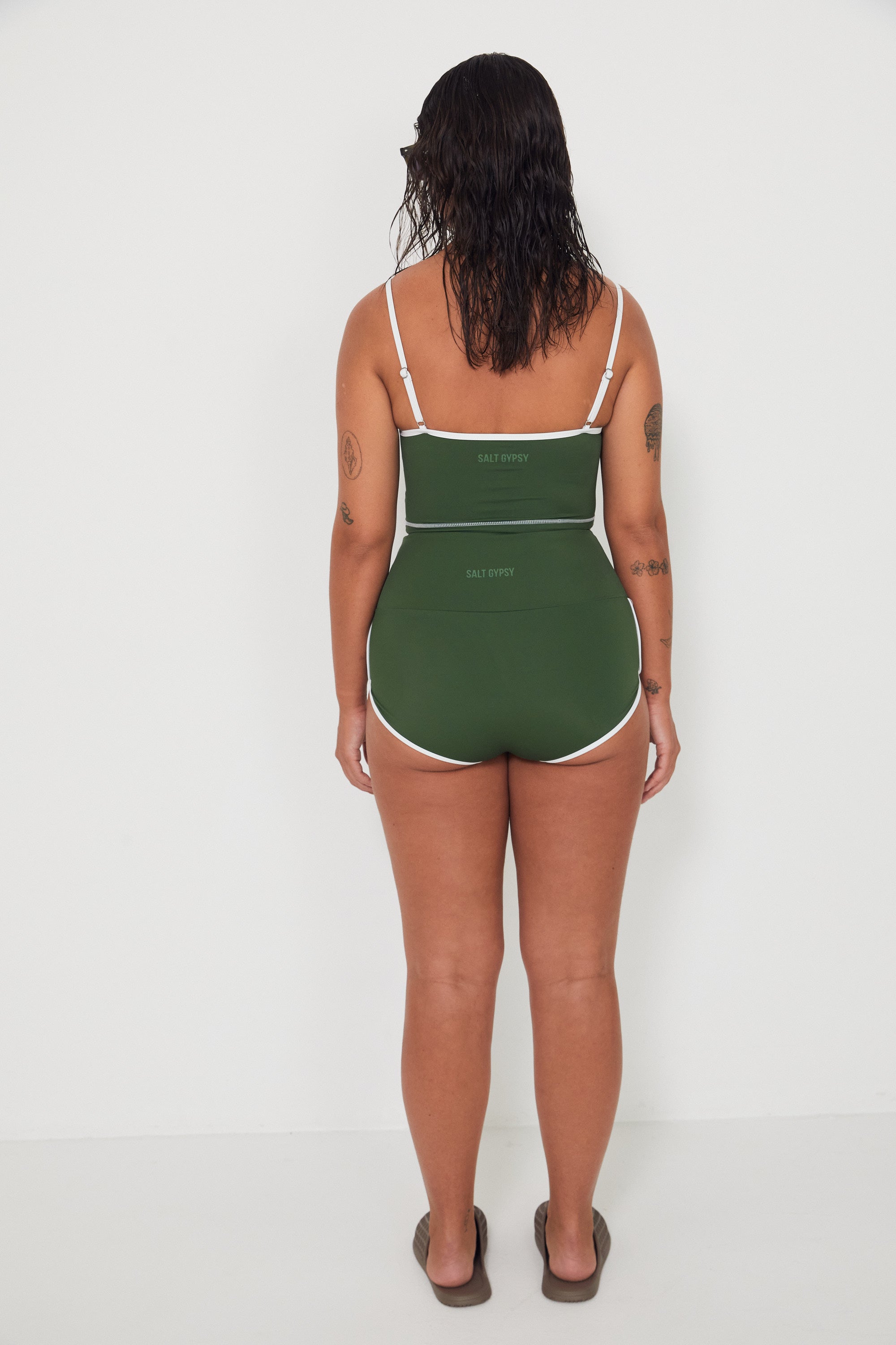 Woman wearing a green tankini surf top and betty bottoms 2.0 swimsuit with white trim from the back against a white background