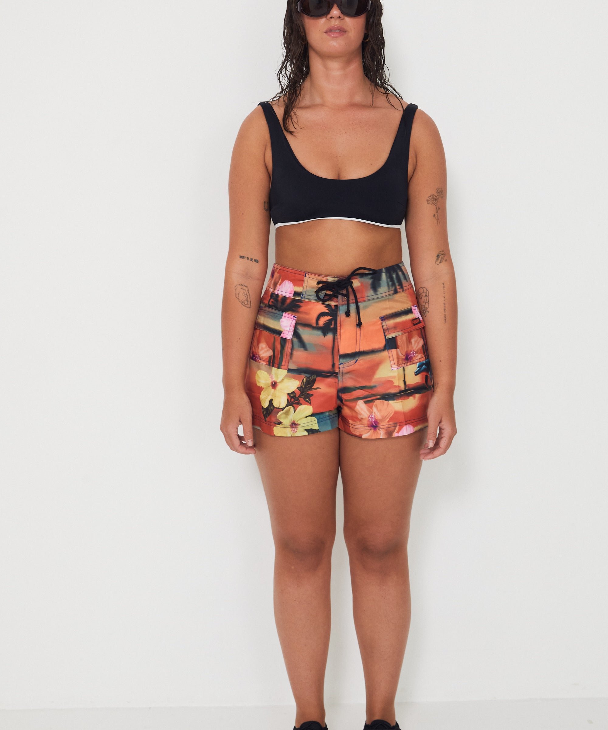 Woman wearing a black bralette swimwear crop top and vacay floral holiday swimwear boardshorts on a white background collaboration of salt gypsy and friday 