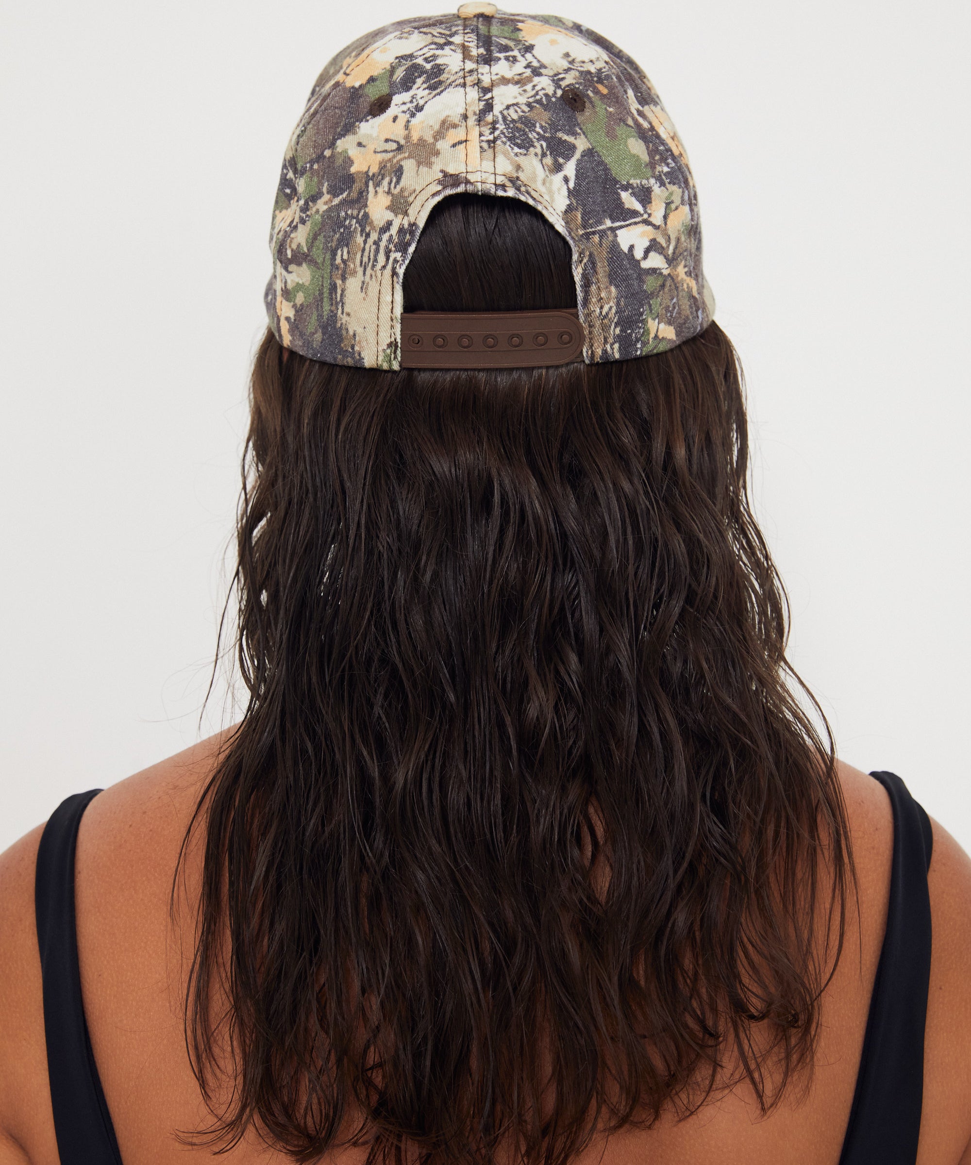 Person wearing a camouflage dad cap with long beachy hair flowing out of it on a white background